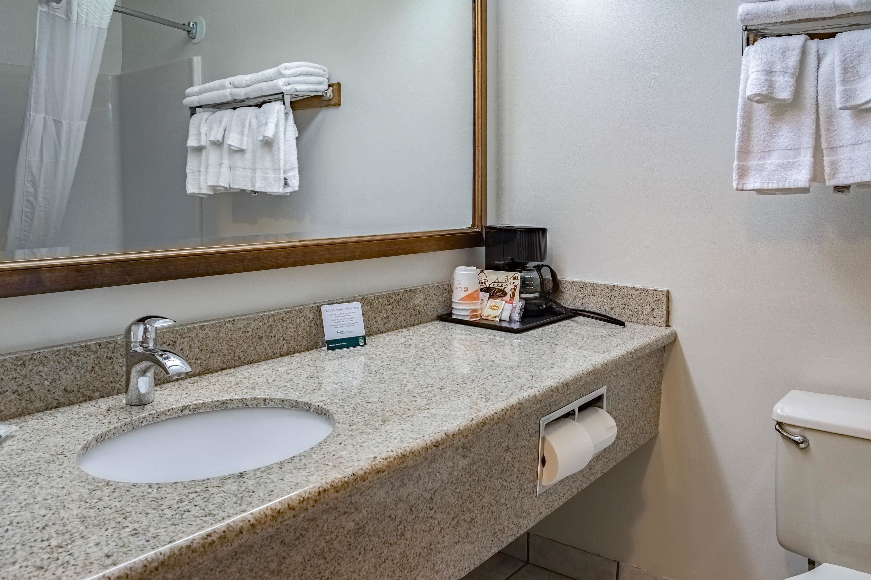A bathroom with a granite countertop sink, a mirror, a towel rack with folded towels, a coffee maker, and a toilet paper holder.