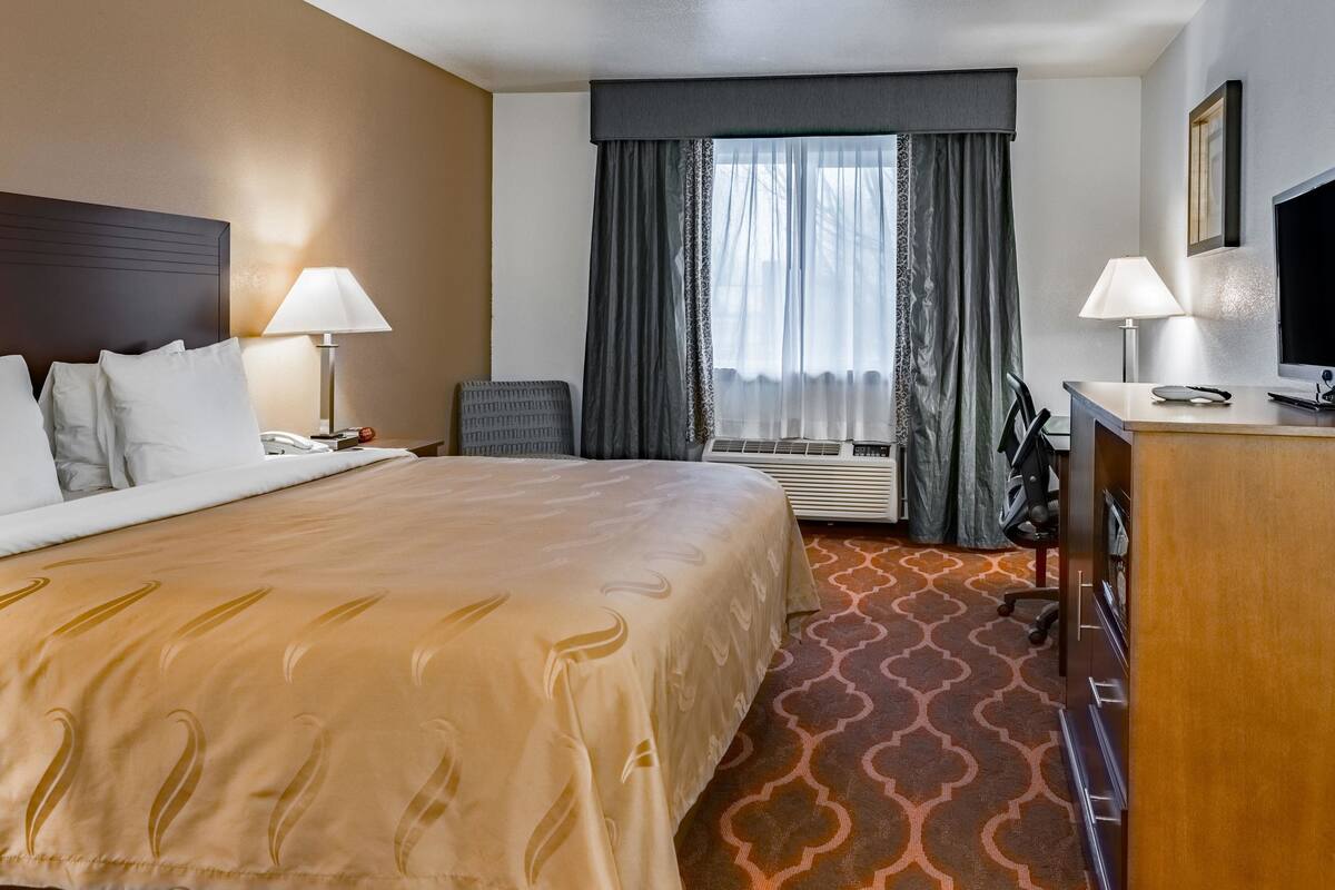 A hotel room with a large bed, two bedside lamps, a desk with a computer, a chair, and a large window with curtains.