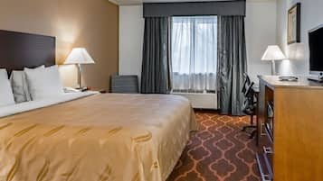 A hotel room with a large bed, two bedside lamps, a desk with a computer, a chair, and a large window with curtains.