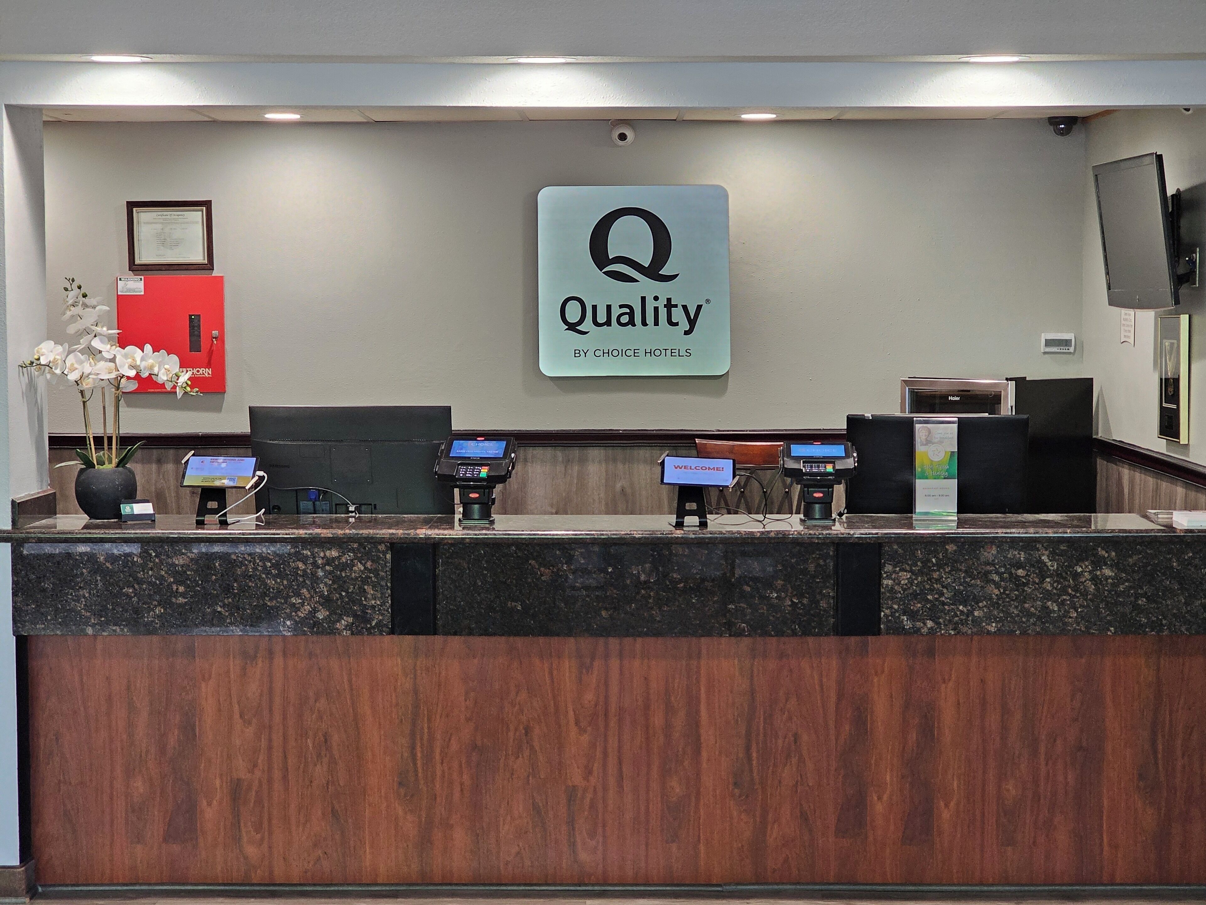 A hotel reception desk with a 'Quality by Choice Hotels' sign, a red flower arrangement, and three payment terminals.
