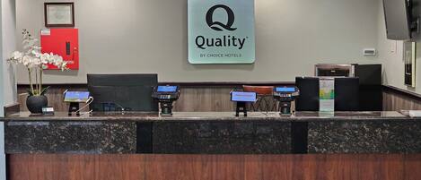 A hotel reception desk with a 'Quality by Choice Hotels' sign, a red flower arrangement, and three payment terminals.