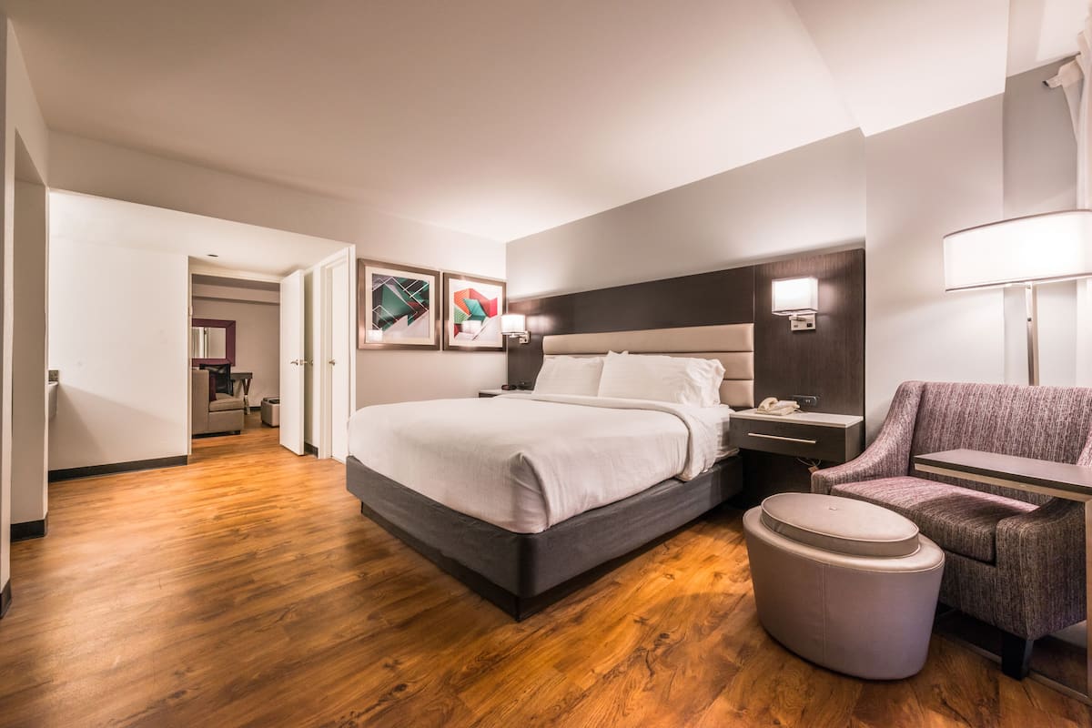 A modern hotel room with a large bed, a seating area with a chair, a small table, and a nightstand with a lamp.