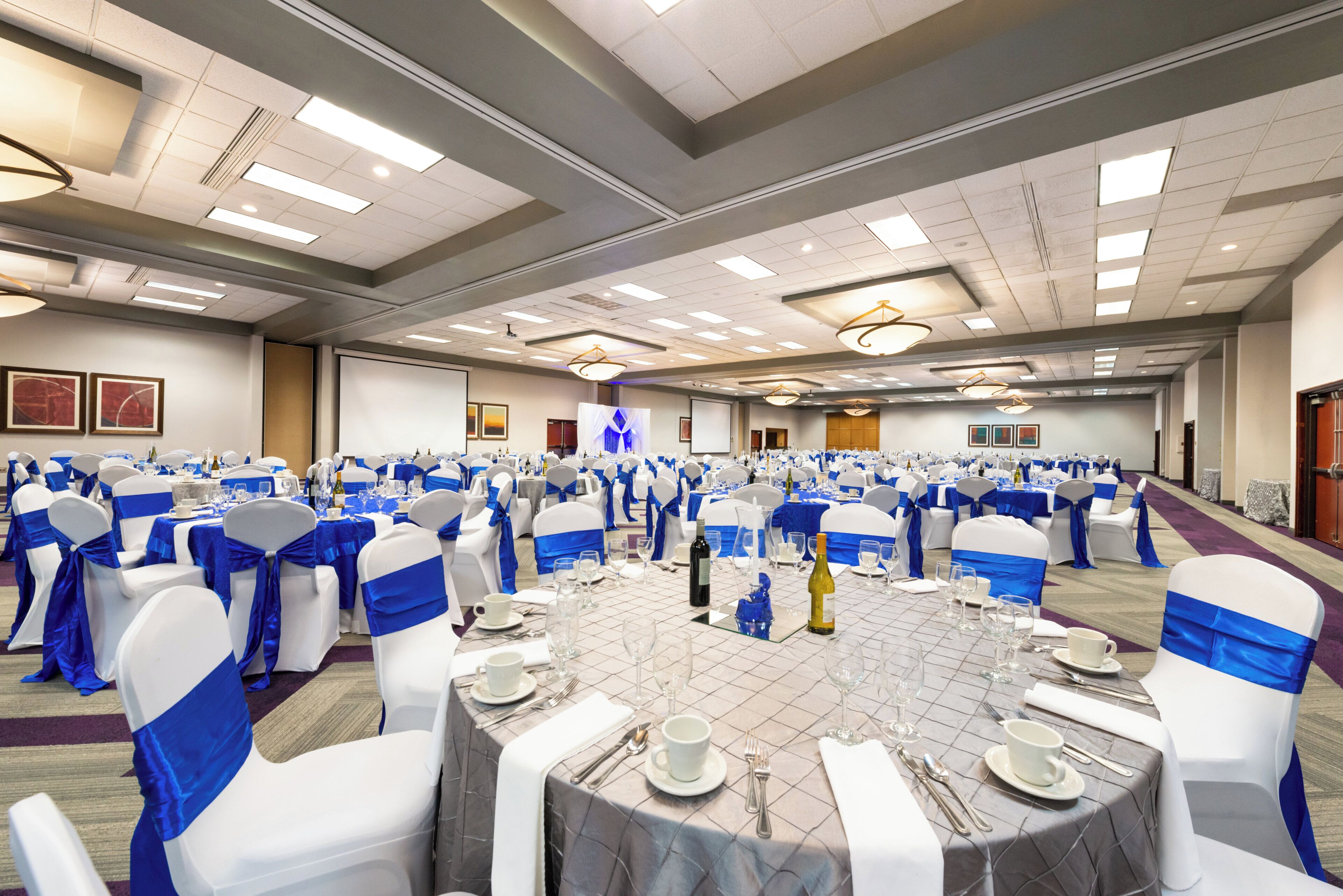 Un salón de banquetes con mesas redondas dispuestas para cenar, sillas con cintas azules e iluminación empotrada en el techo.