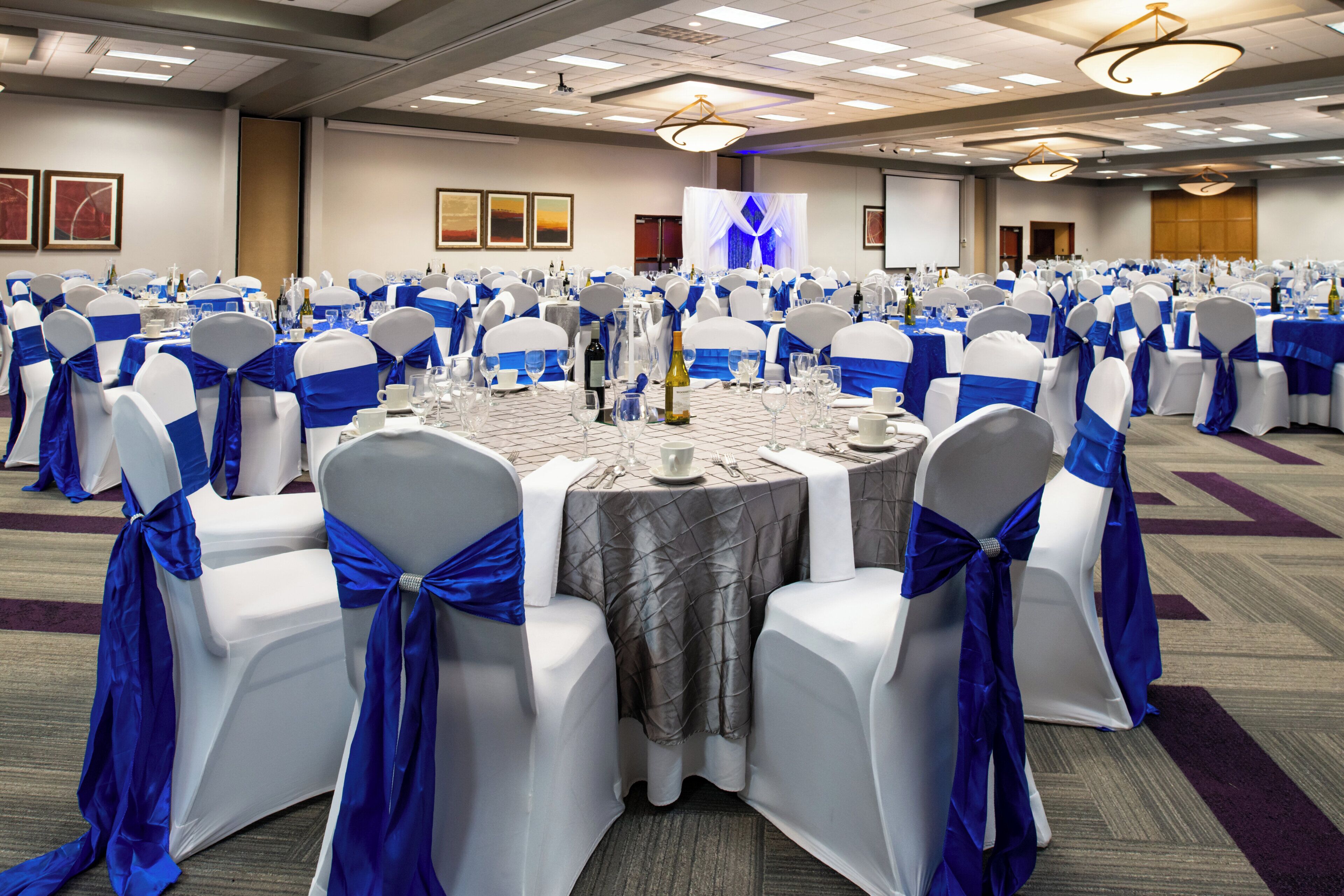 A banquet hall with round tables, chairs with blue sashes, and a stage with a blue backdrop.
