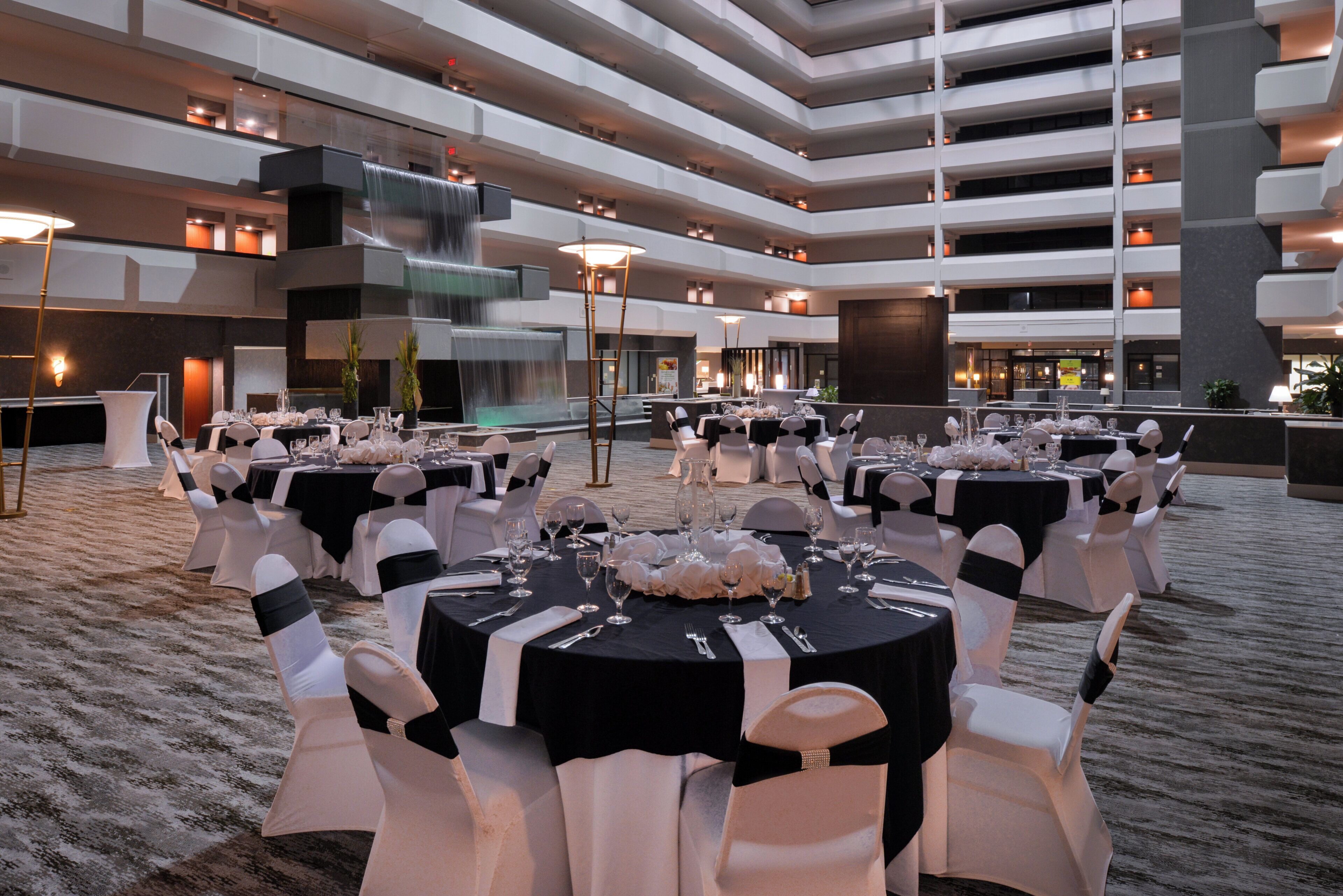 A multi-level indoor event space with round tables set for a formal occasion, featuring a central water feature and modern architecture.