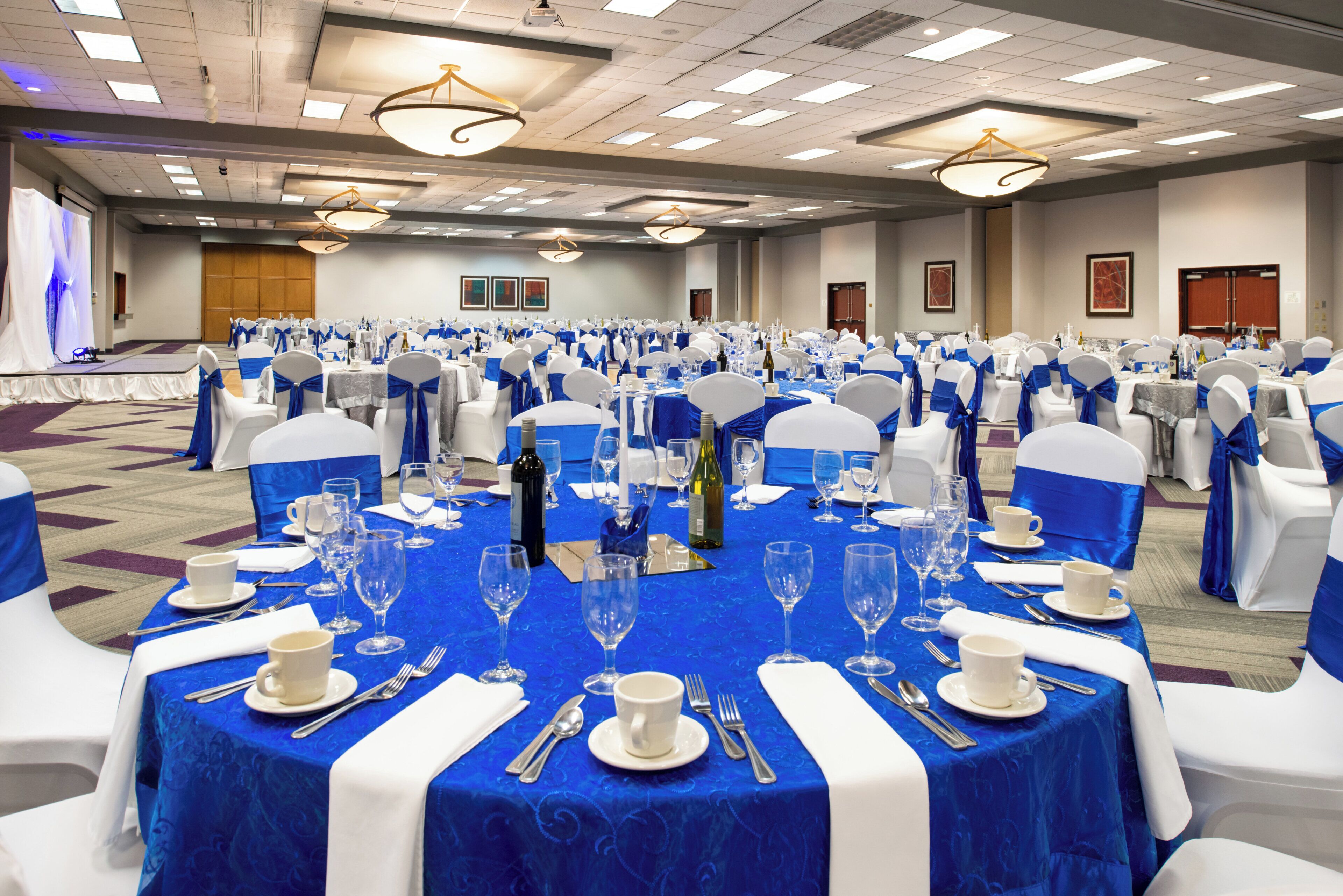 A banquet hall with round tables set for a formal event, featuring white chairs with blue sashes, and tables covered with blue tablecloths.