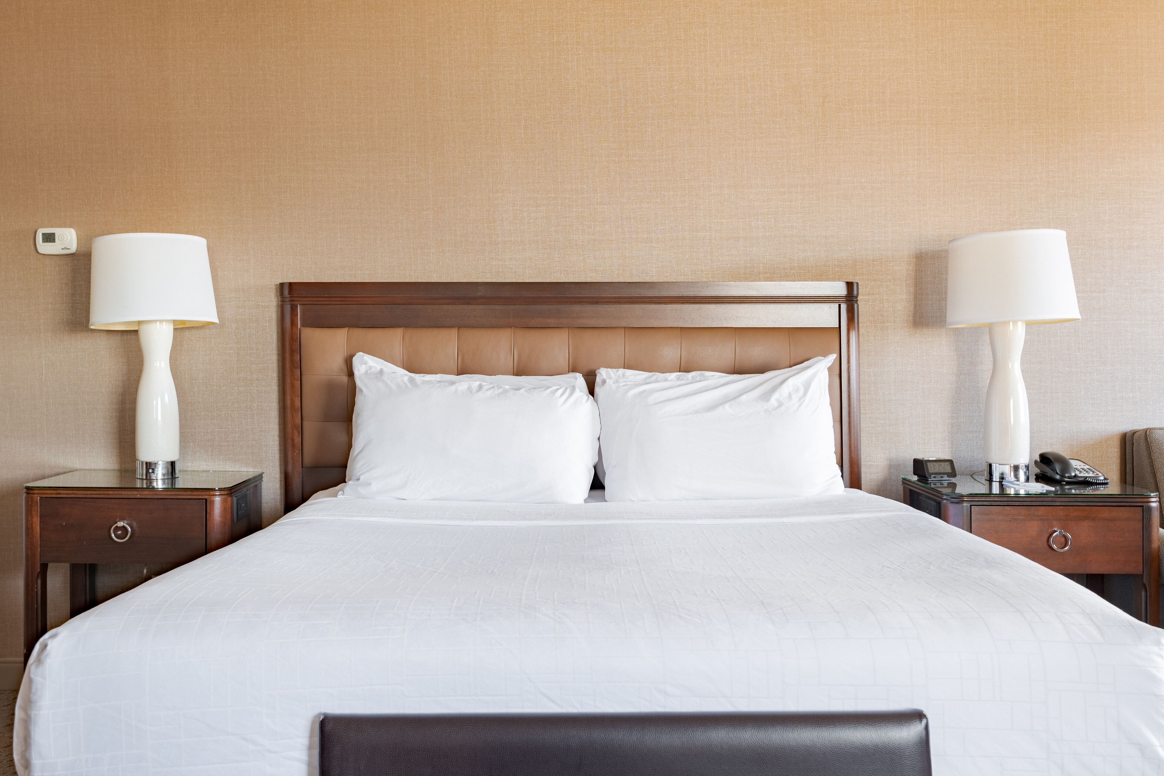 Habitación de hotel con una cama grande, dos mesitas de noche con lámparas, una mesita auxiliar con teléfono y un termostato fijado a la pared.