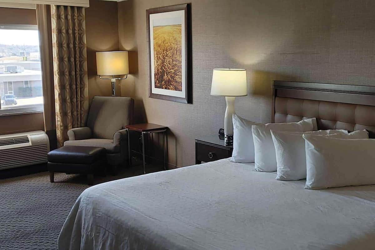 A hotel room with a bed, two bedside lamps, a chair, a small table, a picture on the wall, and an air conditioning unit.
