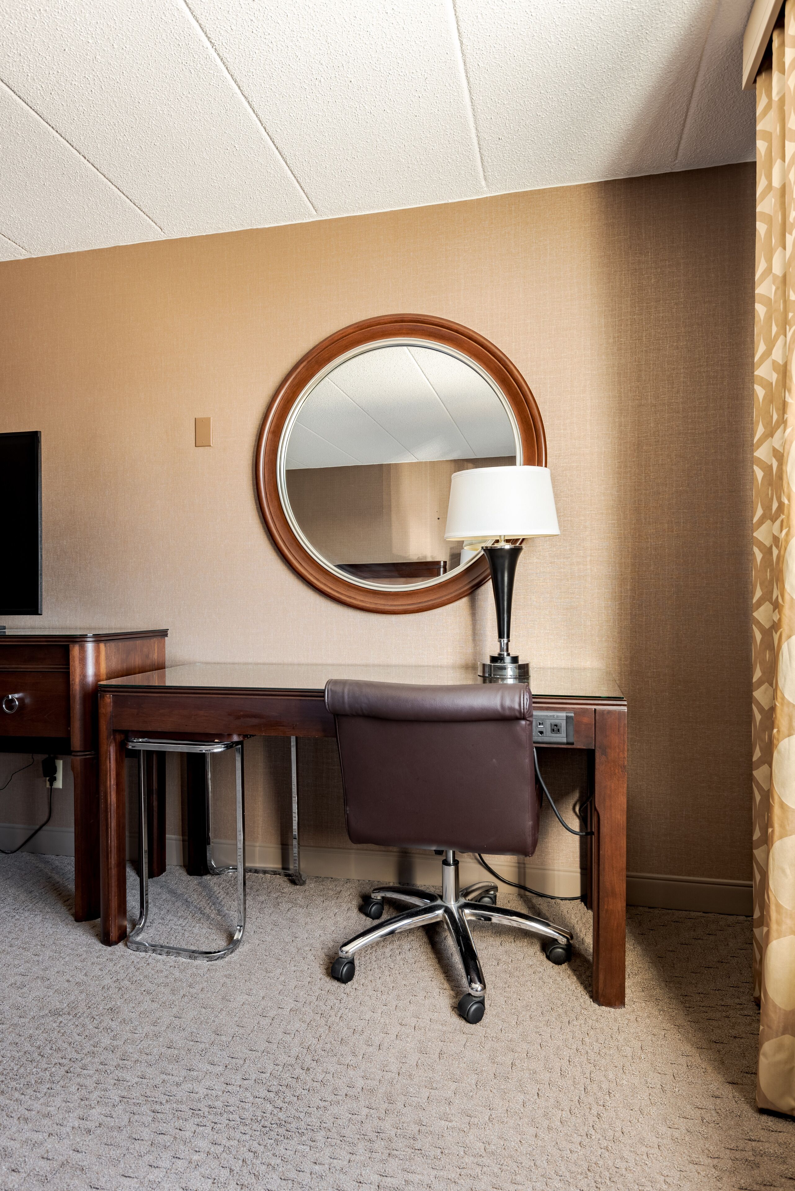 Une chambre d’hôtel avec un bureau en bois, une chaise, un miroir rond et une lampe.