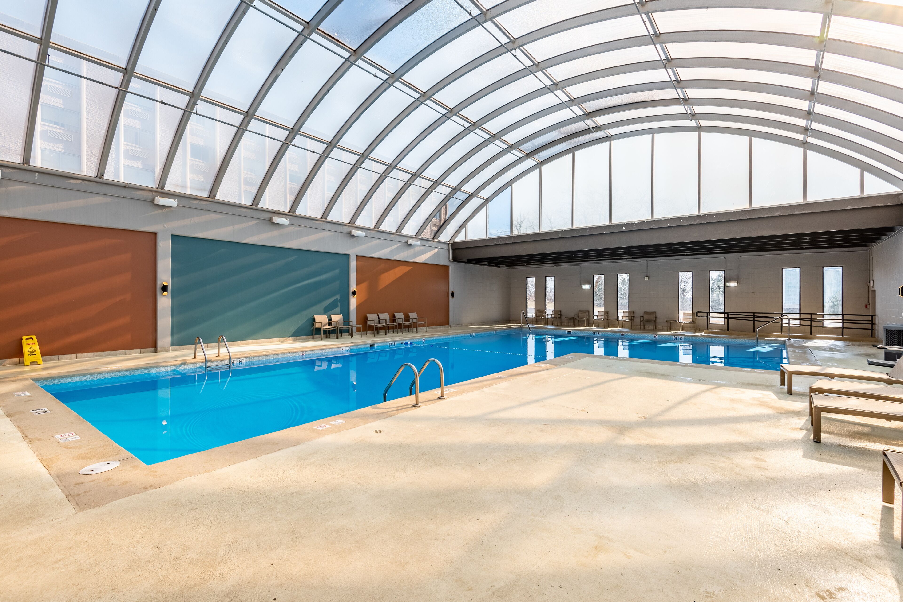 An indoor swimming pool with a glass ceiling, clear poolside chairs, and a lifeguard chair.