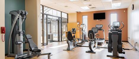 A gym with various exercise machines, a large window, and a television on the wall.