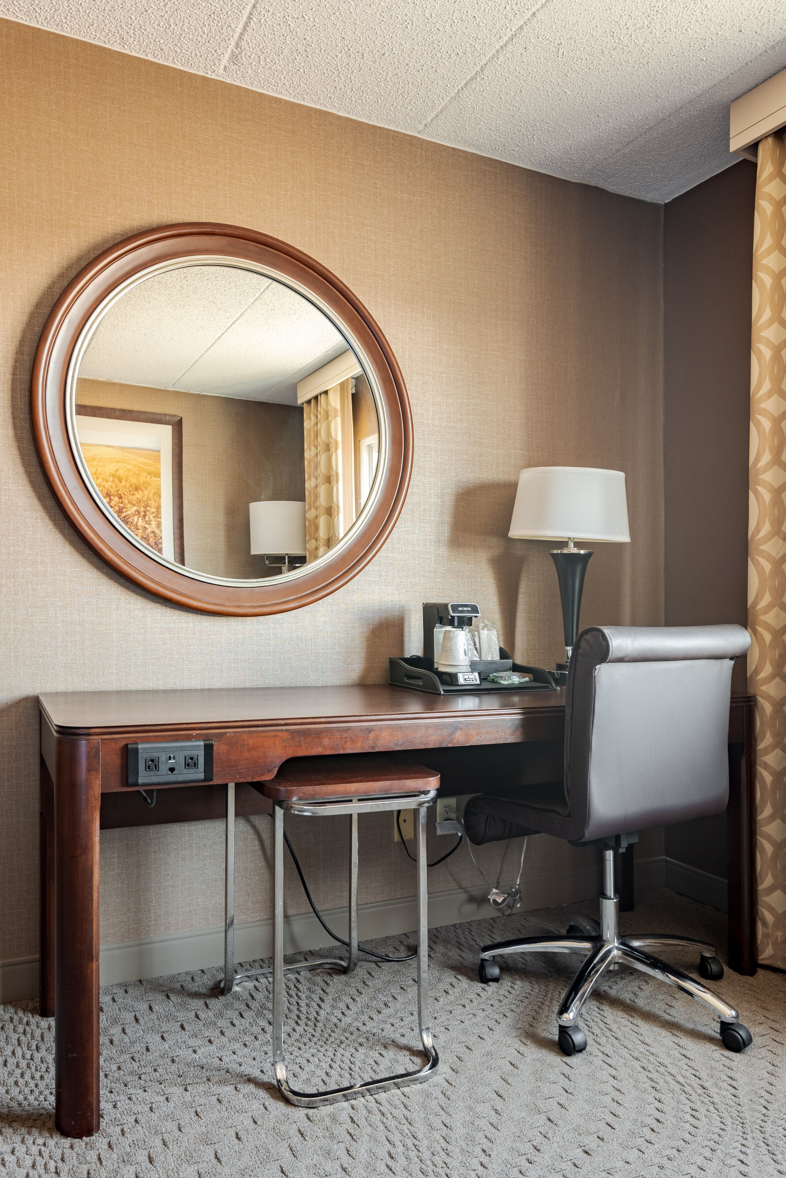 A hotel room with a wooden desk, a modern chair, a round mirror, a lamp, and a small table with a coffee maker.