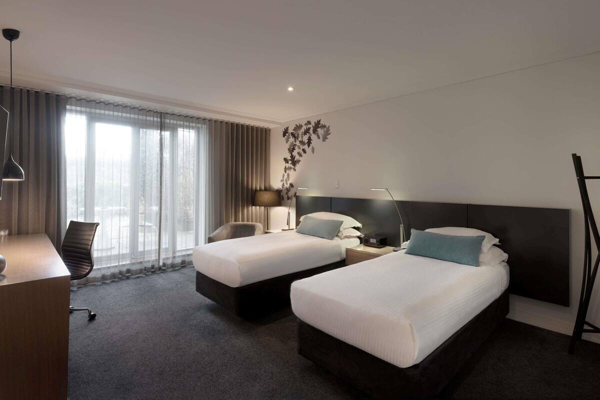 A hotel room with two beds, a desk, a chair, a lamp, and a large window.