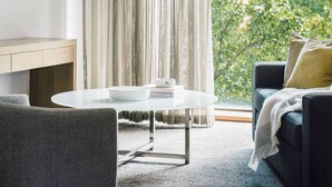 A modern living room with a round white table, a dark sofa with a throw blanket, and a view of greenery outside. - The Como Melbourne by Accor (South Yarra)