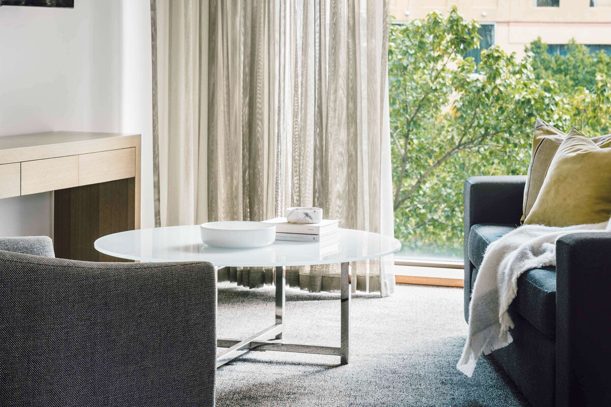 A modern living room with a round white table, a dark sofa with a throw blanket, and a view of greenery outside.