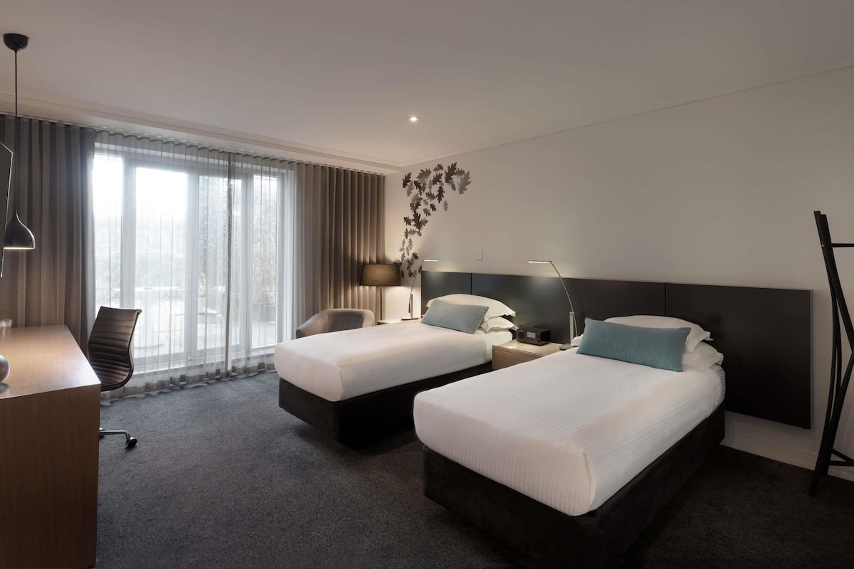 A hotel room with two beds, a desk, a chair, a lamp, and a view of the outdoors.