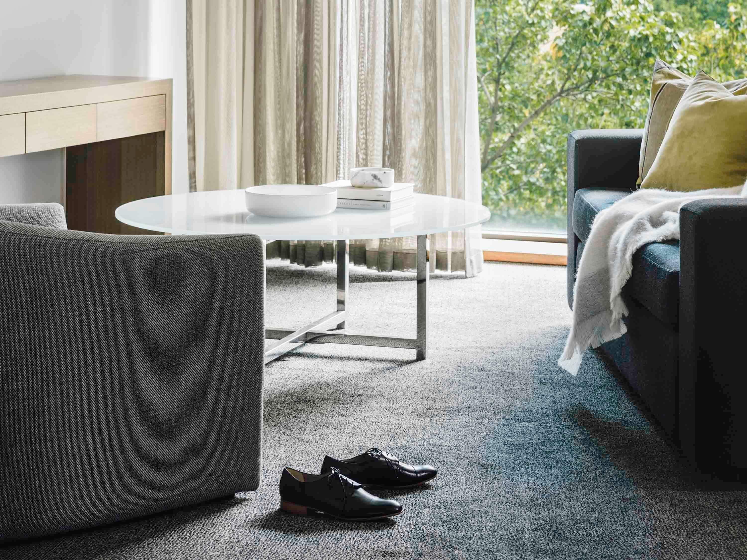 Un salon moderne avec une table basse ronde en verre, un canapé gris, un fauteuil noir et une paire de chaussures posées sur le sol.