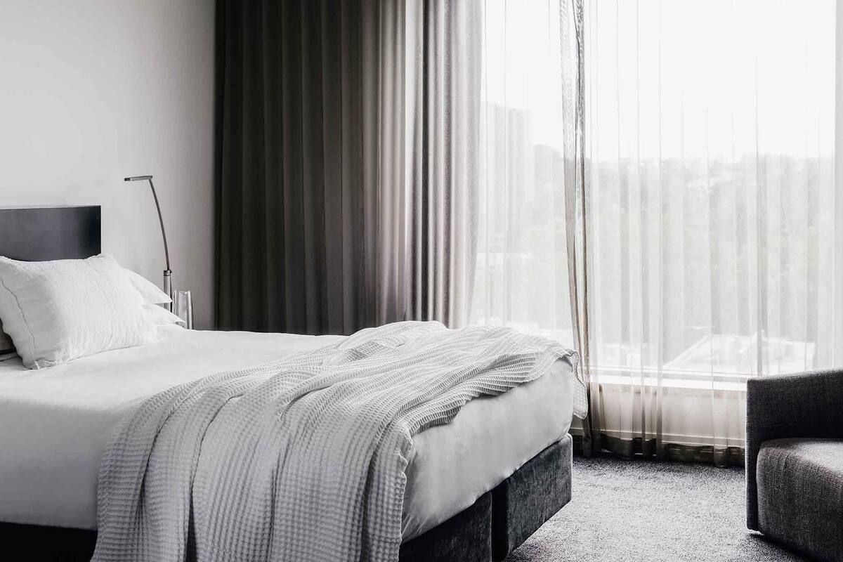 A hotel room with a bed, a chair, a lamp, and a view of the city through sheer curtains.
