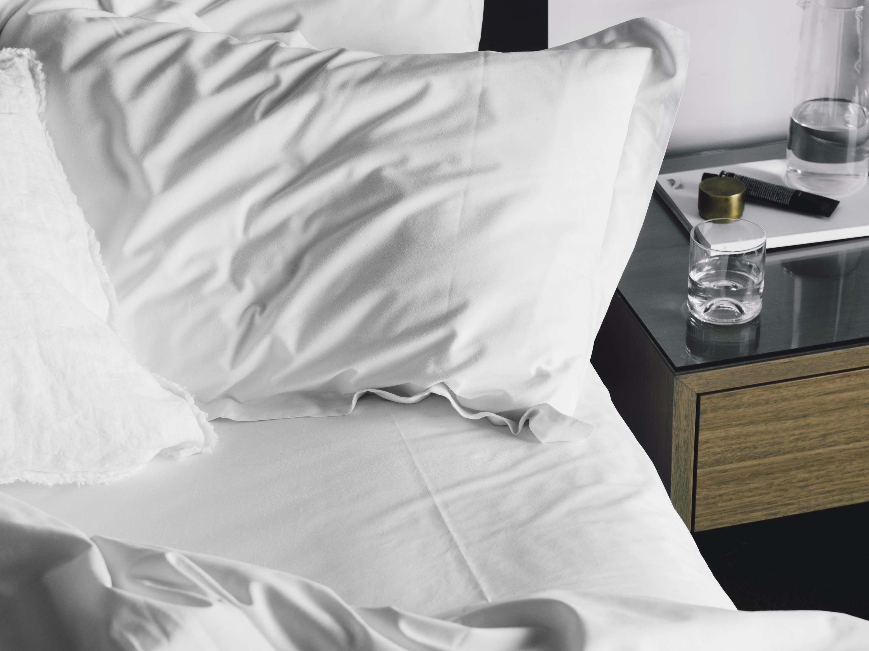 Una cama bien hecha con sábanas blancas, una mesita de noche con un vaso de agua, una botella pequeña y un control remoto.