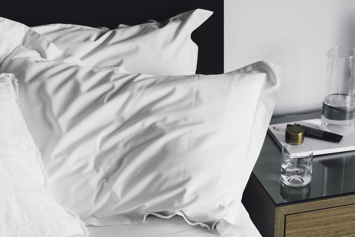 A neatly made bed with white linens, a black nightstand with a glass of water, and a small black object on the nightstand.