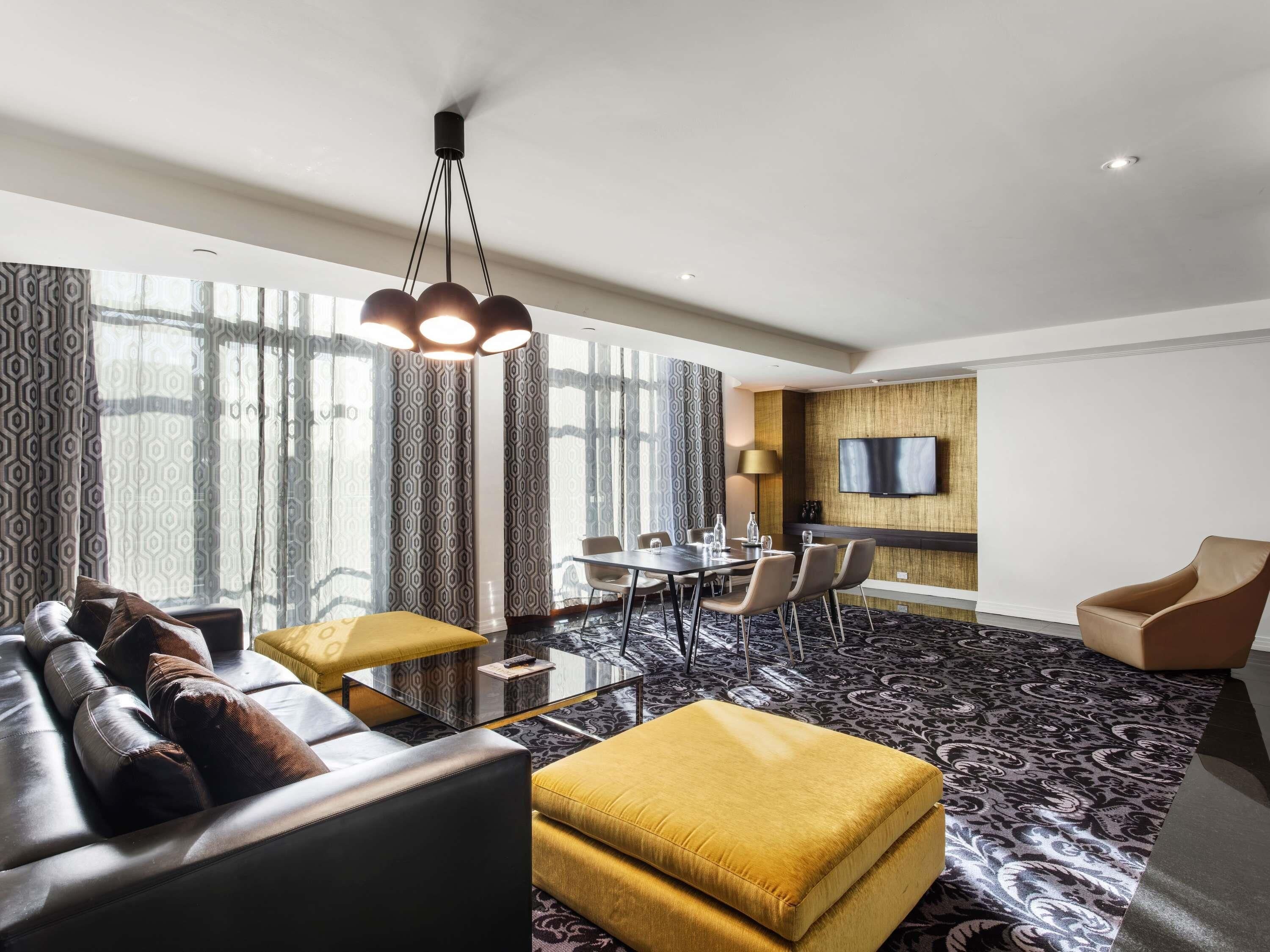 A modern hotel room with a sofa, ottoman, dining table, chairs, and a TV mounted on the wall.