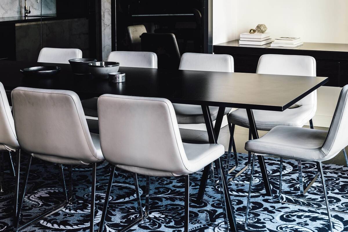 A modern dining area with a black table and white chairs, a patterned carpet, and a kitchen in the background.