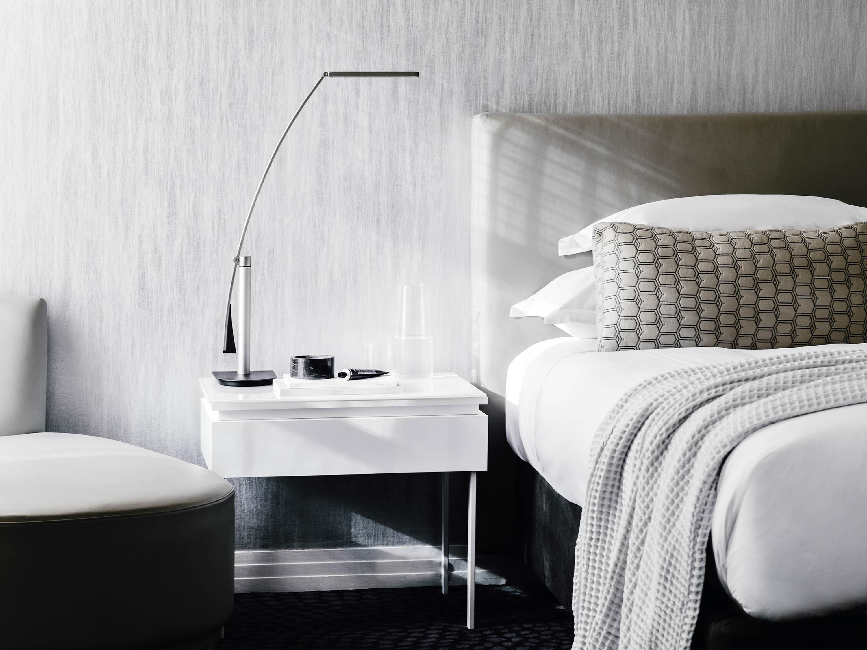 A modern hotel room with a white bedside table, a lamp, a bed with patterned pillows, and a view of the outdoors.