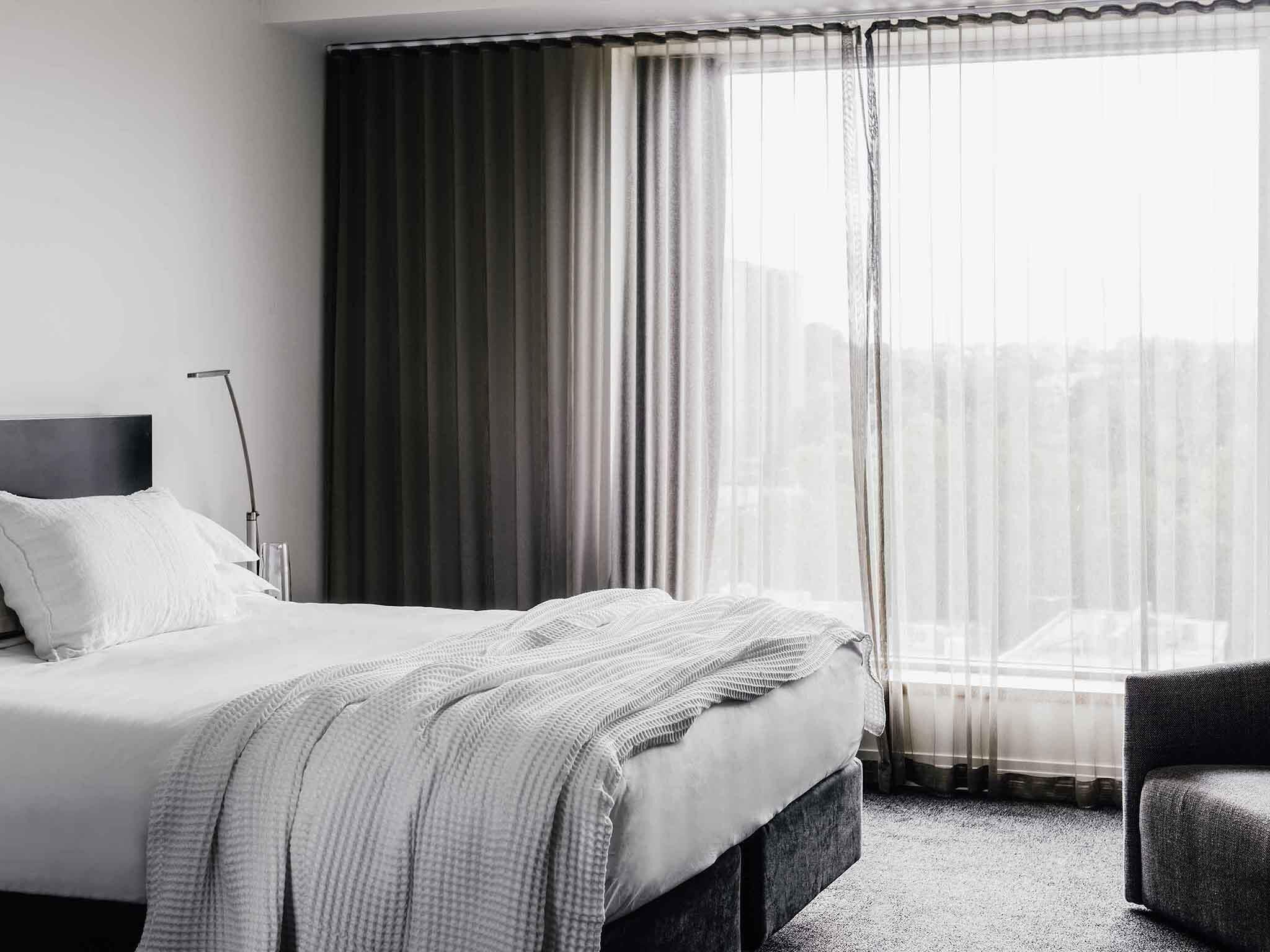 Habitación de hotel con cama, una silla, una lámpara y vistas de la ciudad a través de cortinas translúcidas.