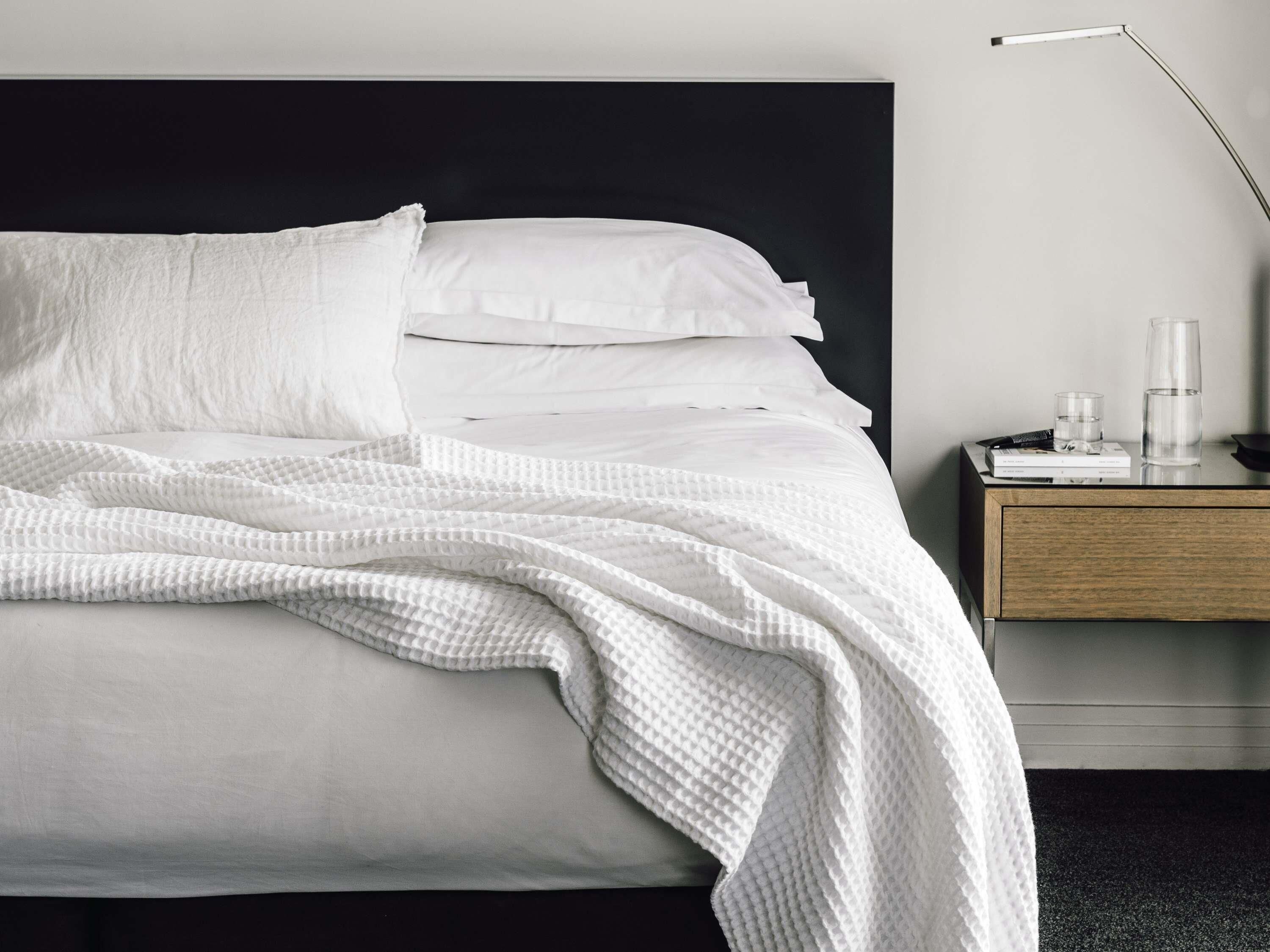 Una cama bien hecha con ropa de cama blanca, un cabecero negro y una mesita de noche con una lámpara, libros y gafas.
