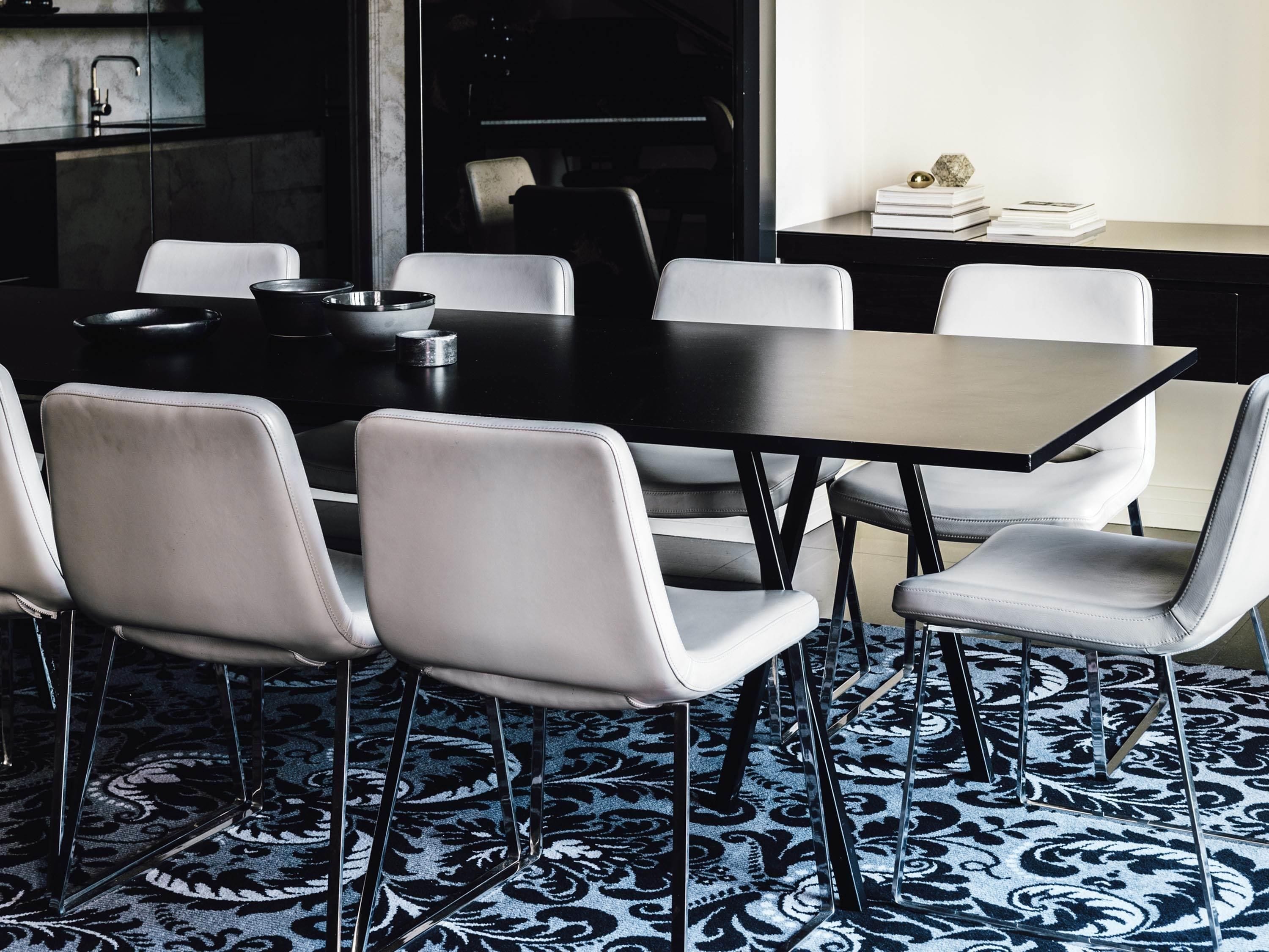 A modern dining area with a black table and white chairs, a patterned carpet, and a kitchen in the background.