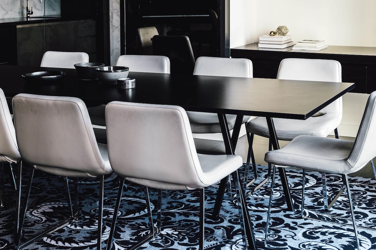 A modern dining area with a black table and white chairs, a patterned carpet, and a kitchen in the background.