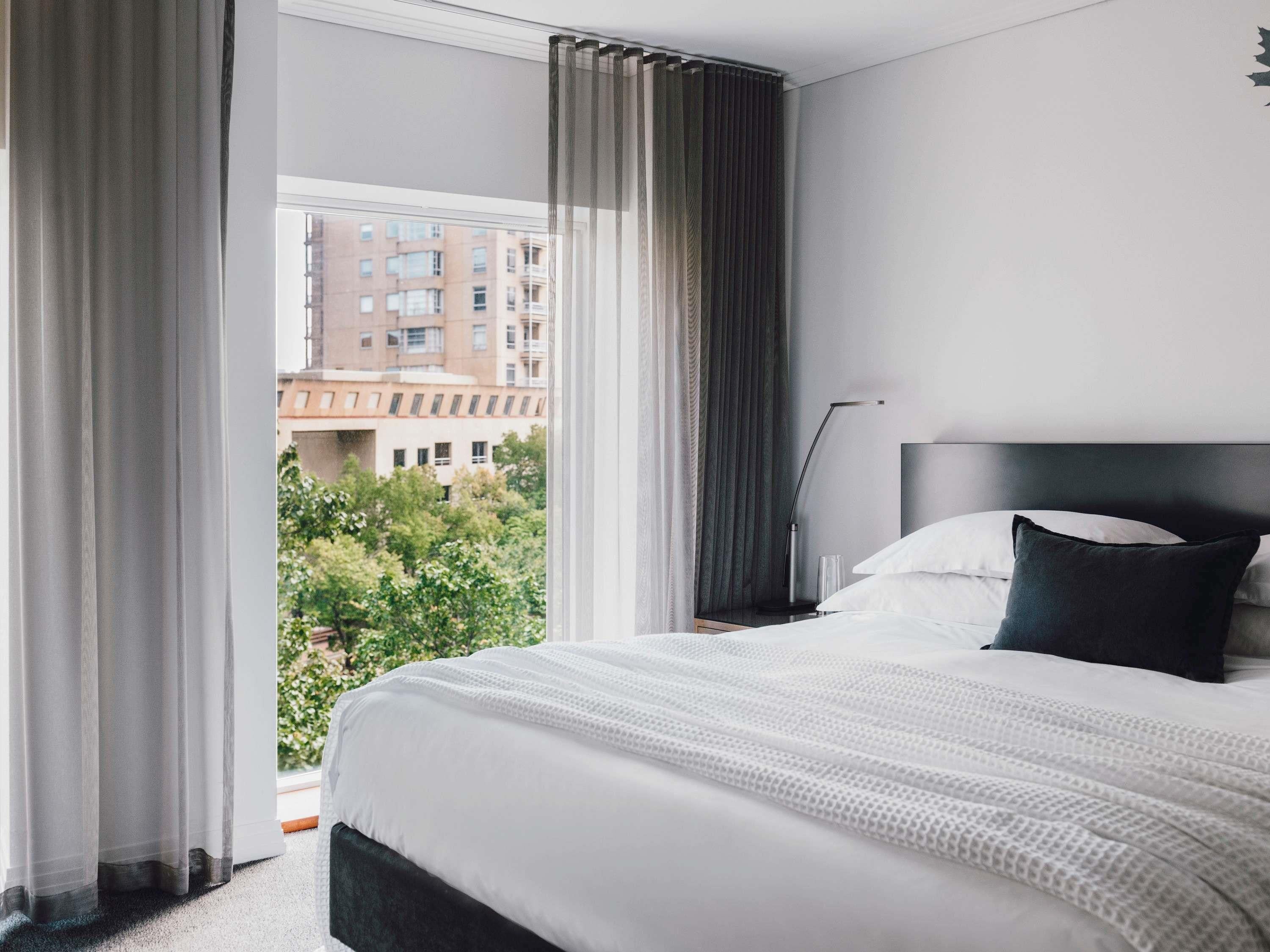 A hotel room with a bed, a nightstand, a lamp, and a view of buildings and trees outside the window.