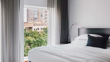 A hotel room with a bed, a nightstand, a lamp, and a view of buildings and trees outside the window.