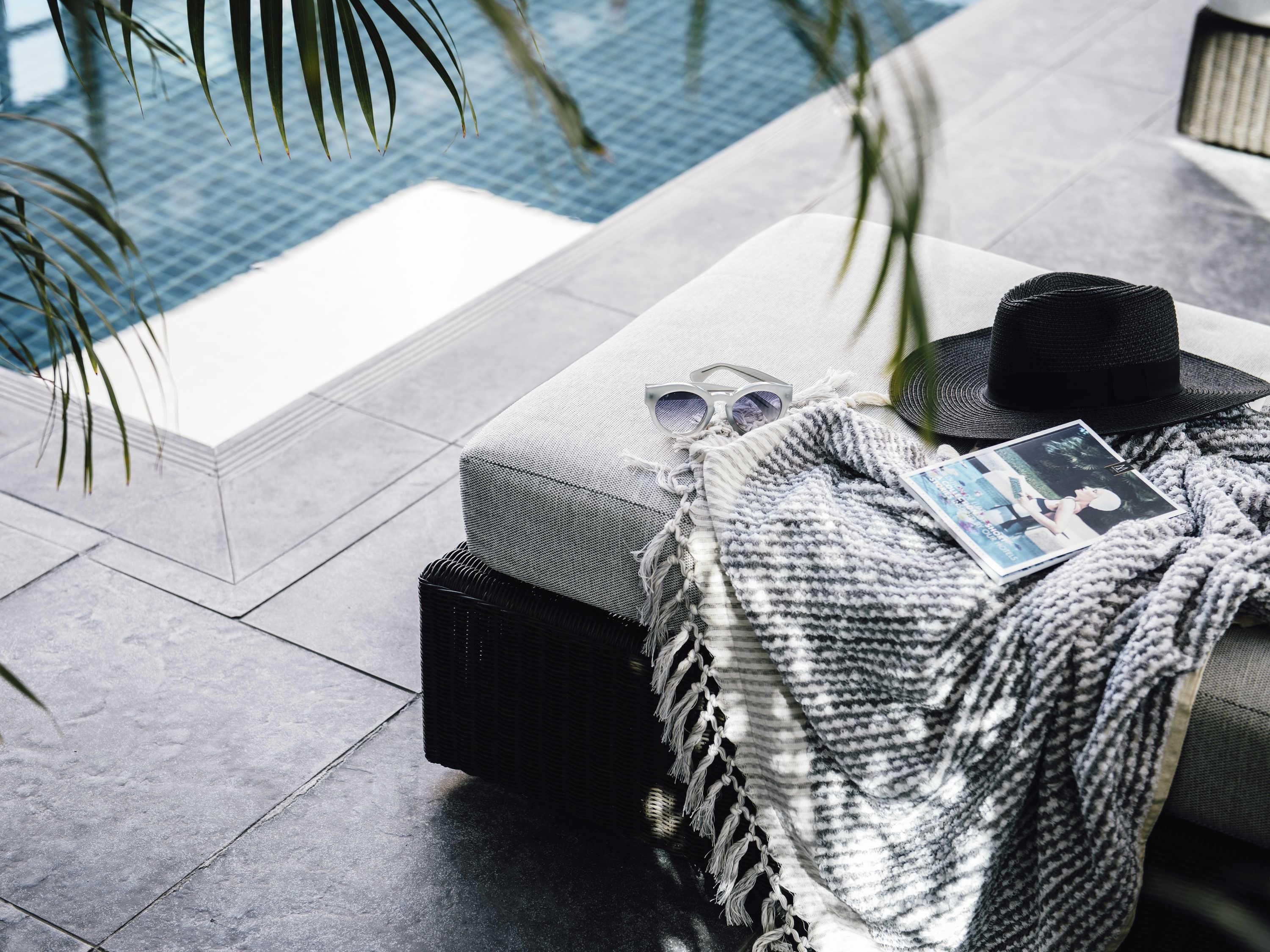A poolside area with a cushioned seating area, a woven blanket, a black hat, and a pair of sunglasses.