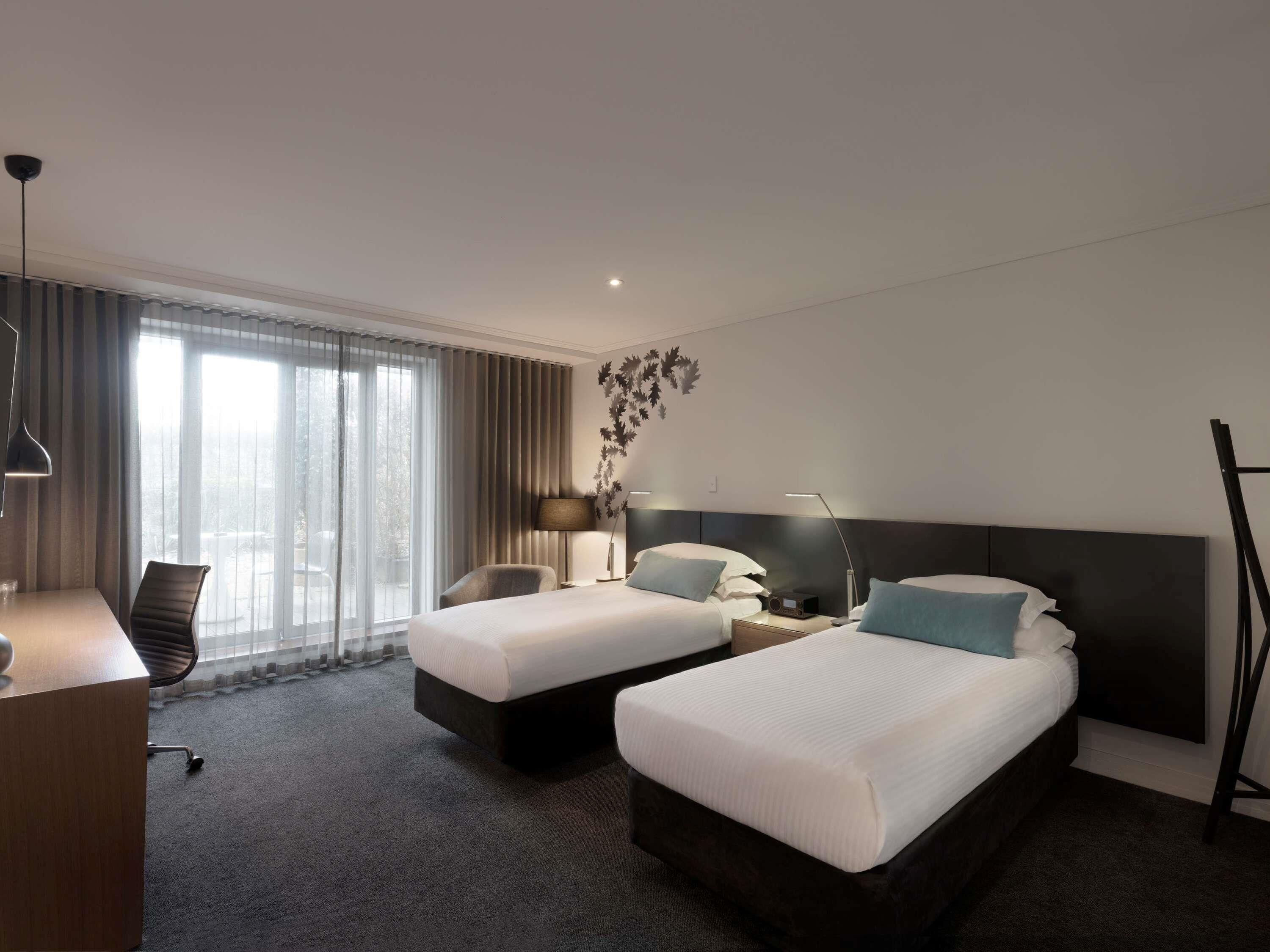 Habitación de hotel con dos camas, un escritorio, una silla, una lámpara y un ventanal grande.