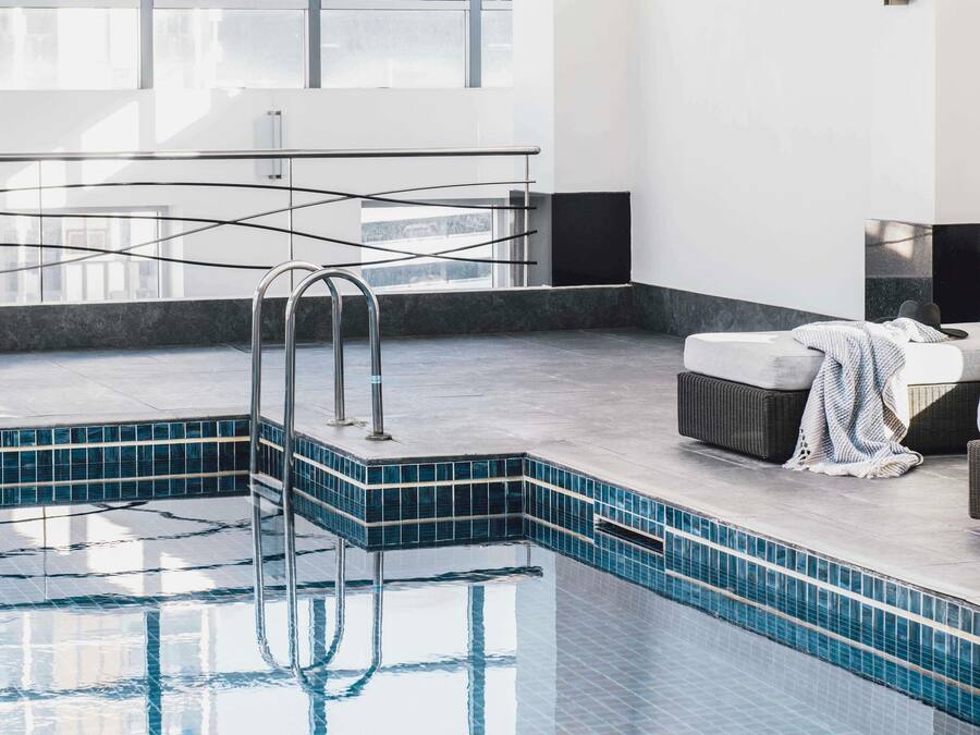 Une piscine intérieure moderne, avec des carreaux bleu clair, une échelle en métal et un espace détente comprenant une chaise en osier et une couverture.