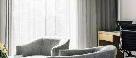 A modern hotel room with two grey armchairs, a small round table, and a wooden desk with a laptop.