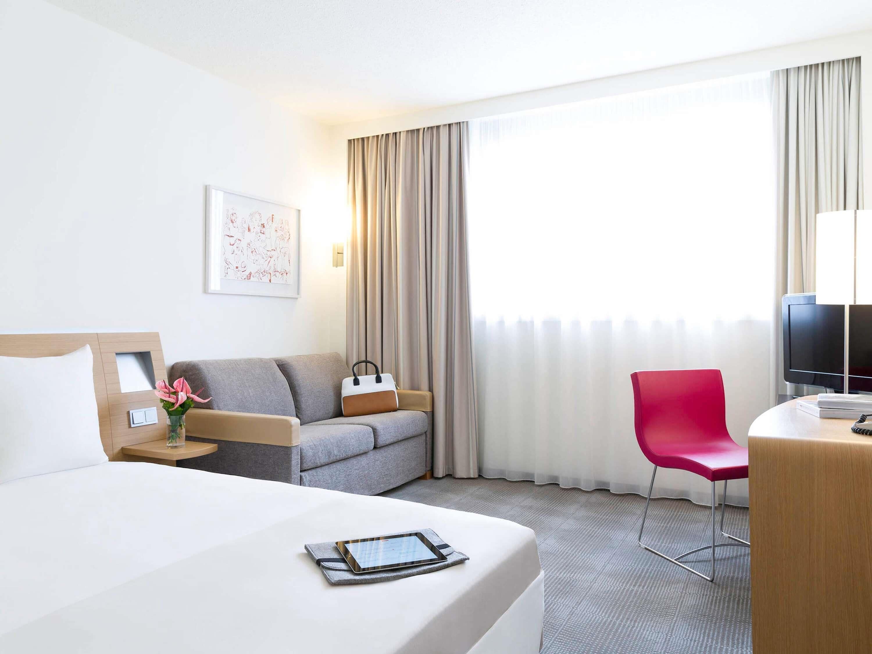A hotel room with a bed, a grey sofa, a red chair, a TV, and a window with curtains.