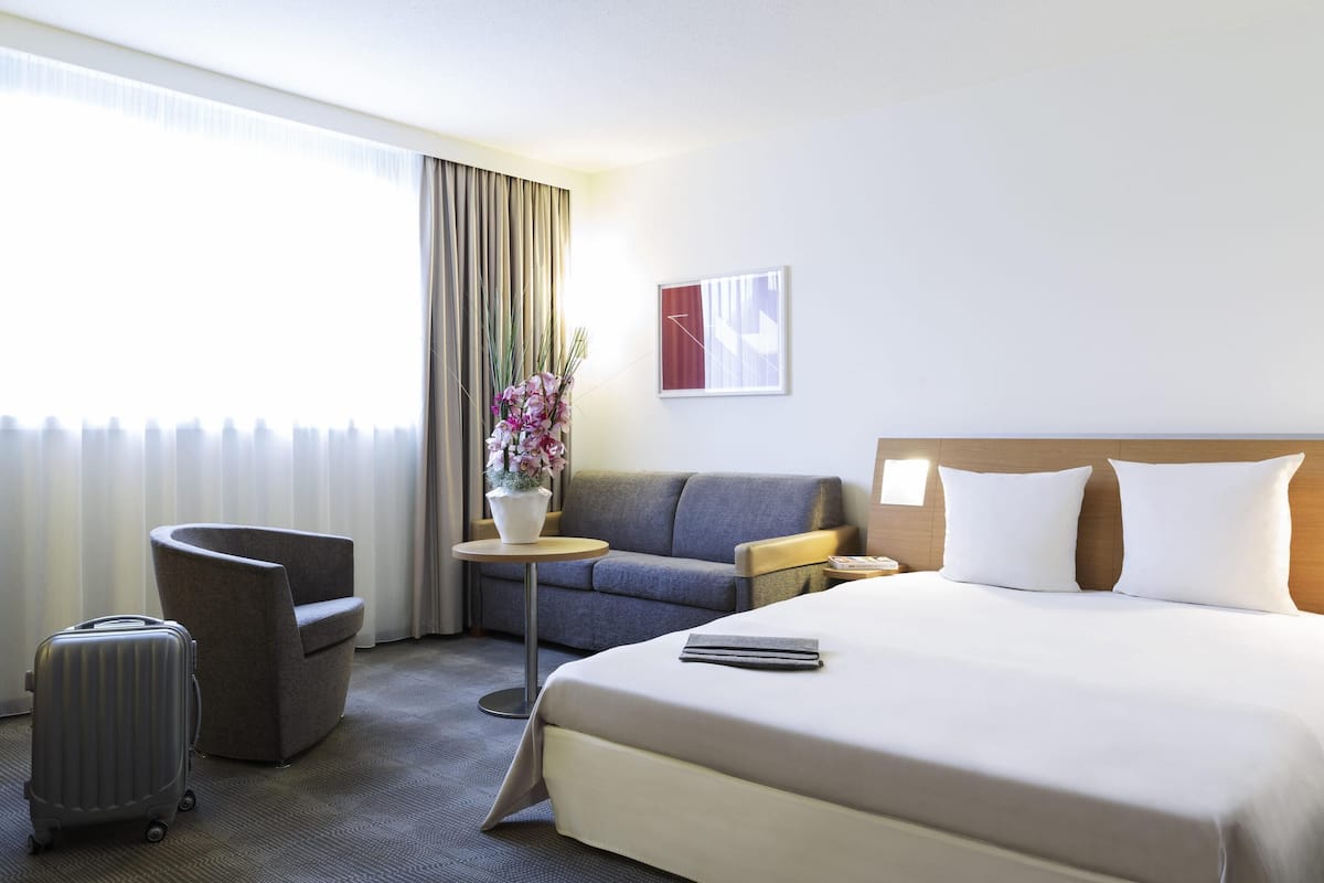 A hotel room with a bed, a sofa, a chair, a small table, a suitcase, and a window with curtains.
