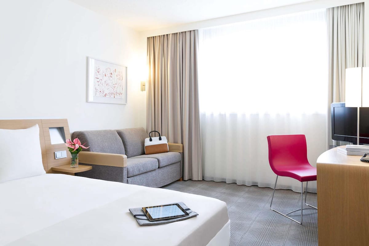 Une chambre d’hôtel avec un lit, un canapé gris, un fauteuil rouge, une télévision et une fenêtre avec des rideaux.
