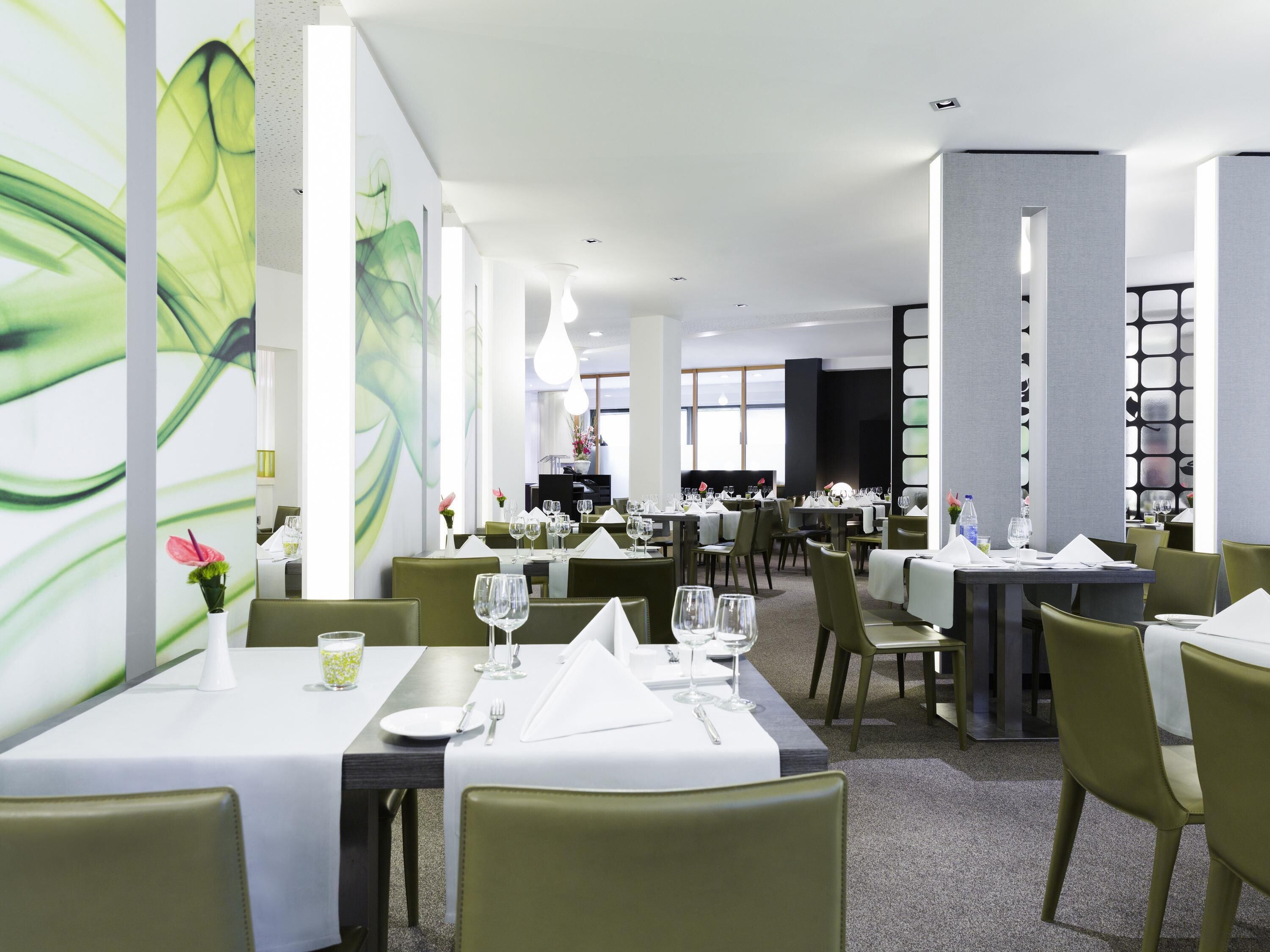 Un restaurant moderne avec des chaises à revêtement vert et des nappes blanches. Les tables sont dressées avec des verres à vin et des couverts. La pièce est dotée d'un haut plafond avec des luminaires encastrés et d'une grande fenêtre qui laisse entrer la lumière naturelle.
