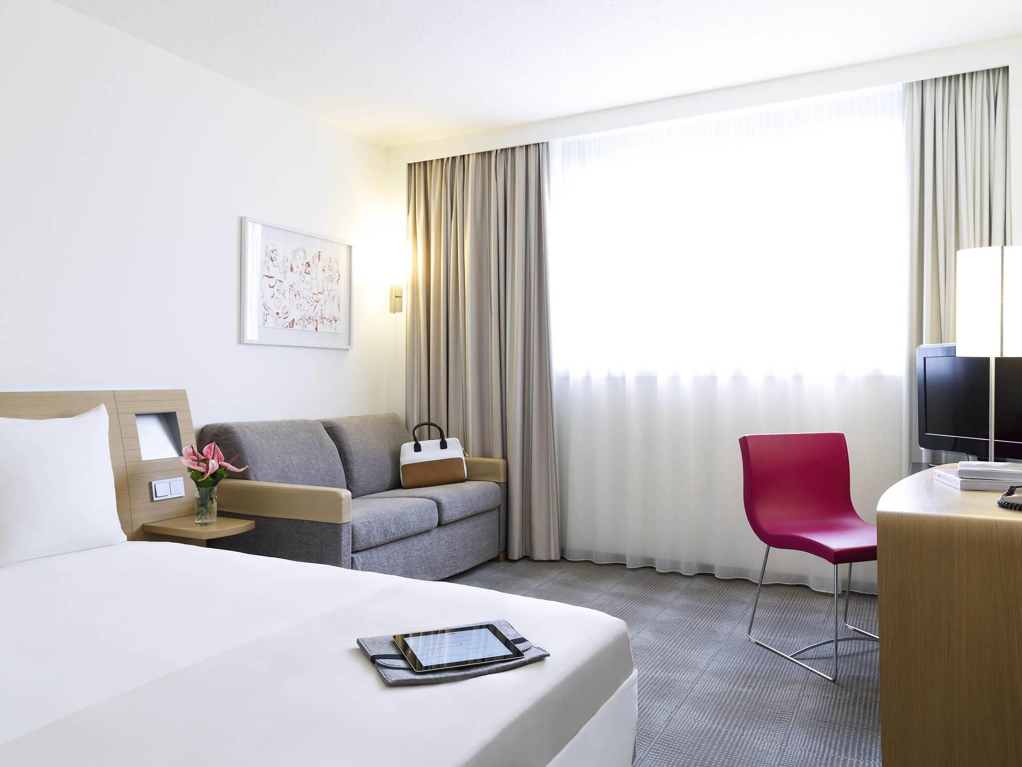 A hotel room with a bed, a grey sofa, a red chair, a desk with a TV, and a window with curtains.
