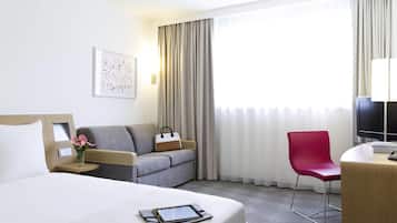 Habitación de hotel con cama, sofá, escritorio con silla, televisor y ventana con cortinas.