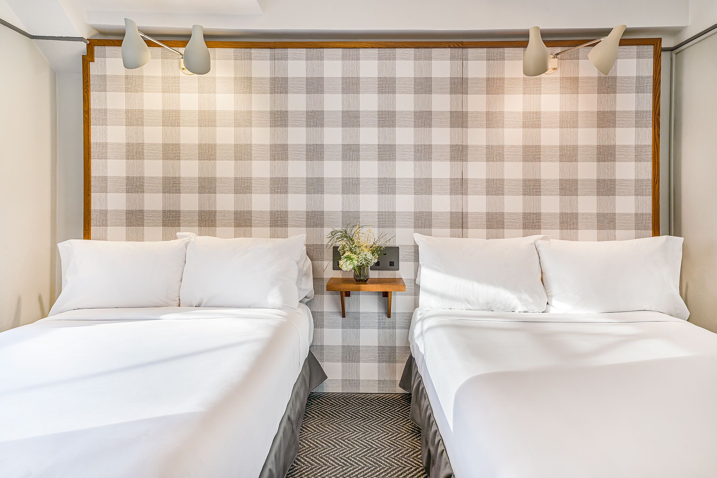 Une chambre d’hôtel avec deux lits, une tête de lit à carreaux, une petite table de chevet ornée d’un vase de fleurs et un parquet à motifs en chevrons.