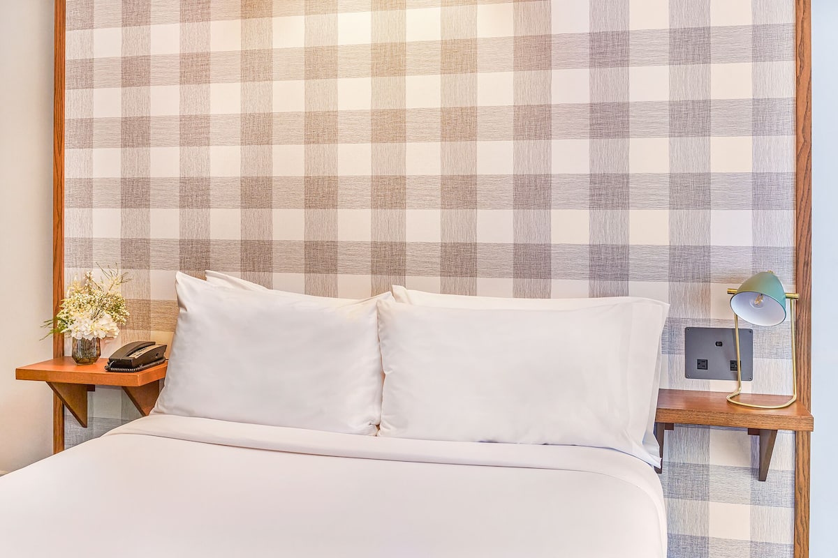 Quarto de hotel com parede quadriculada, cama com roupa de cama branca, mesa de cabeceira com abajur e telefone.