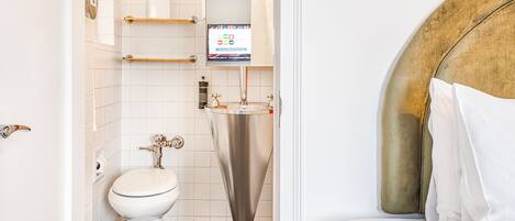 A bathroom with a toilet, a sink, and a shower. There is a digital screen above the sink.