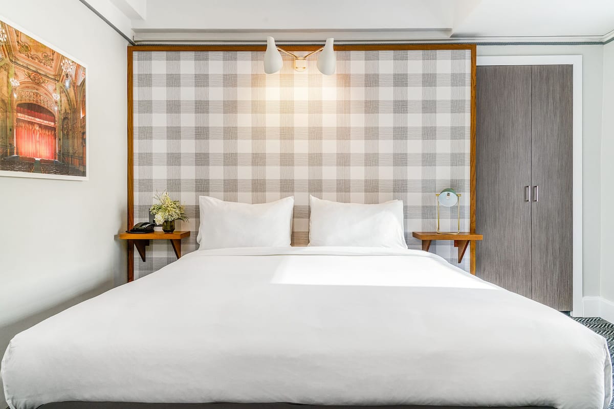 Uma cama bem arrumada com lençóis brancos, cabeceira quadriculada e mesa de cabeceira com abajur e um vaso de flores.