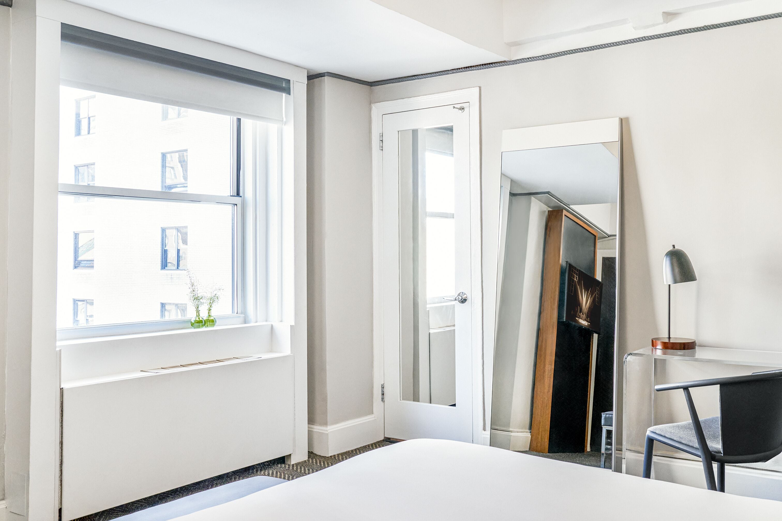 Una camera d'albergo con un letto, una scrivania con una sedia, una finestra vista edifici e una porta.