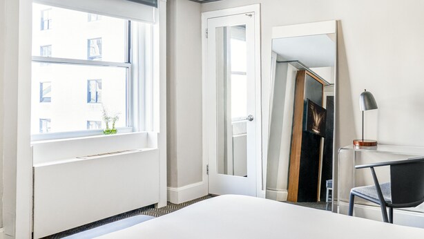 Une chambre d’hôtel comprenant un lit, un bureau avec une chaise, une fenêtre donnant sur des immeubles et une porte.