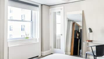 A hotel room with a bed, a desk with a chair, a window with a view of buildings, and a door.